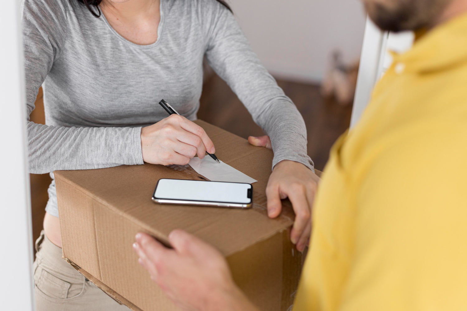 Person signing a document next to a cardboard box with another person handing over a phone.