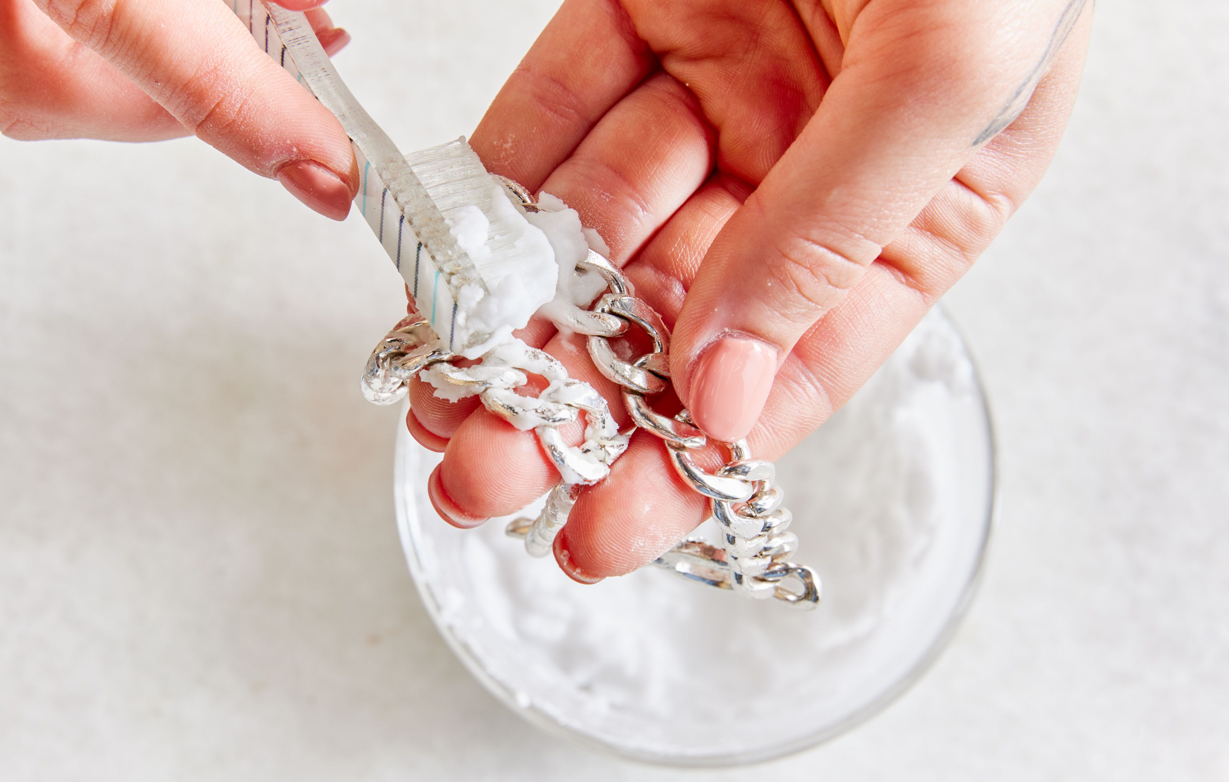 Close-up of hands holding a toothbrush brushing at a silver chain to clean it, with light bakground.