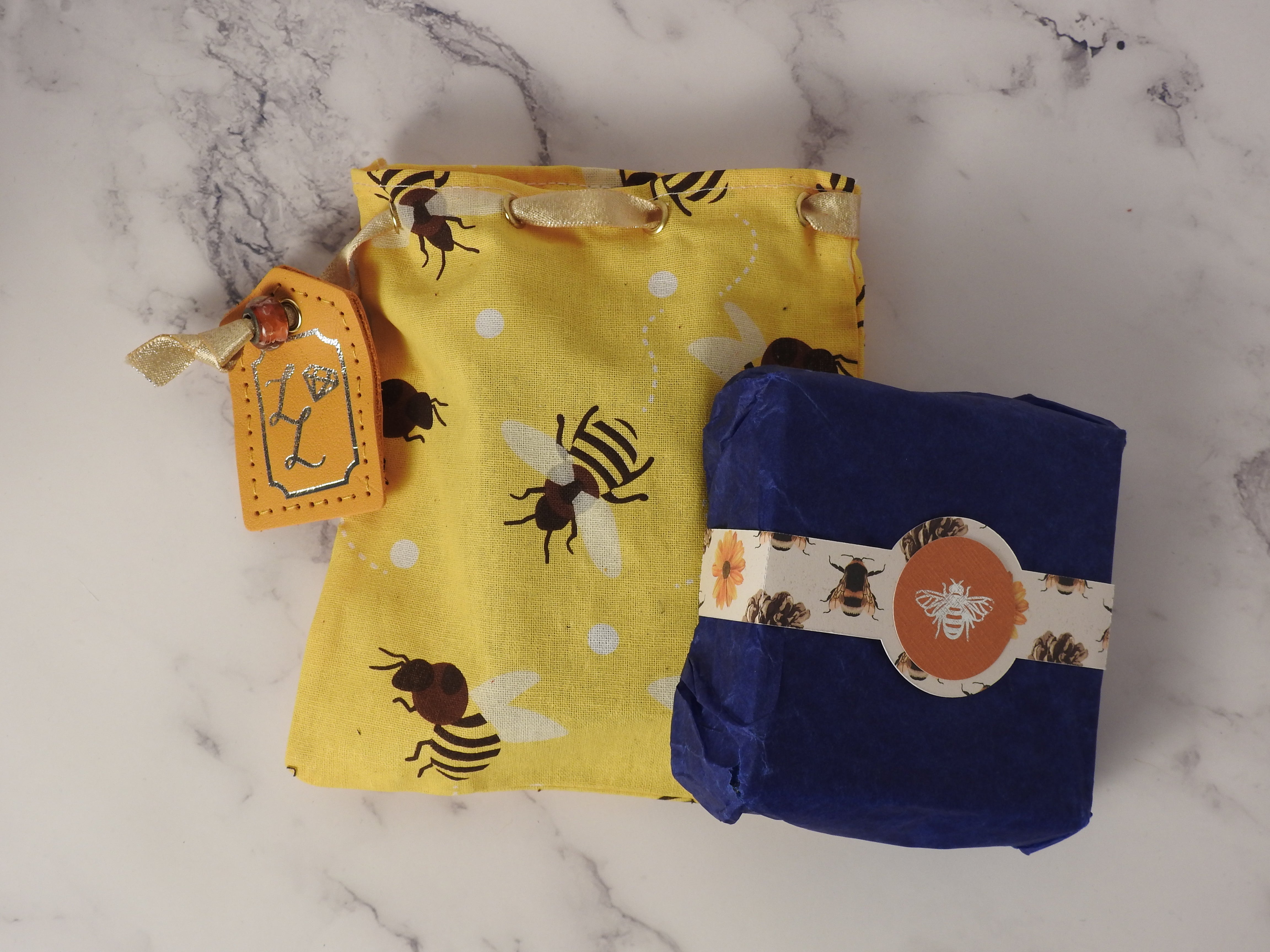close up shot of a cotton bag and cardboard box with belly band. The cotton bag has bee pattern in yellow and a leather tag, in the centre of the bee patterned belly band is a silver foil emblem of a bee.