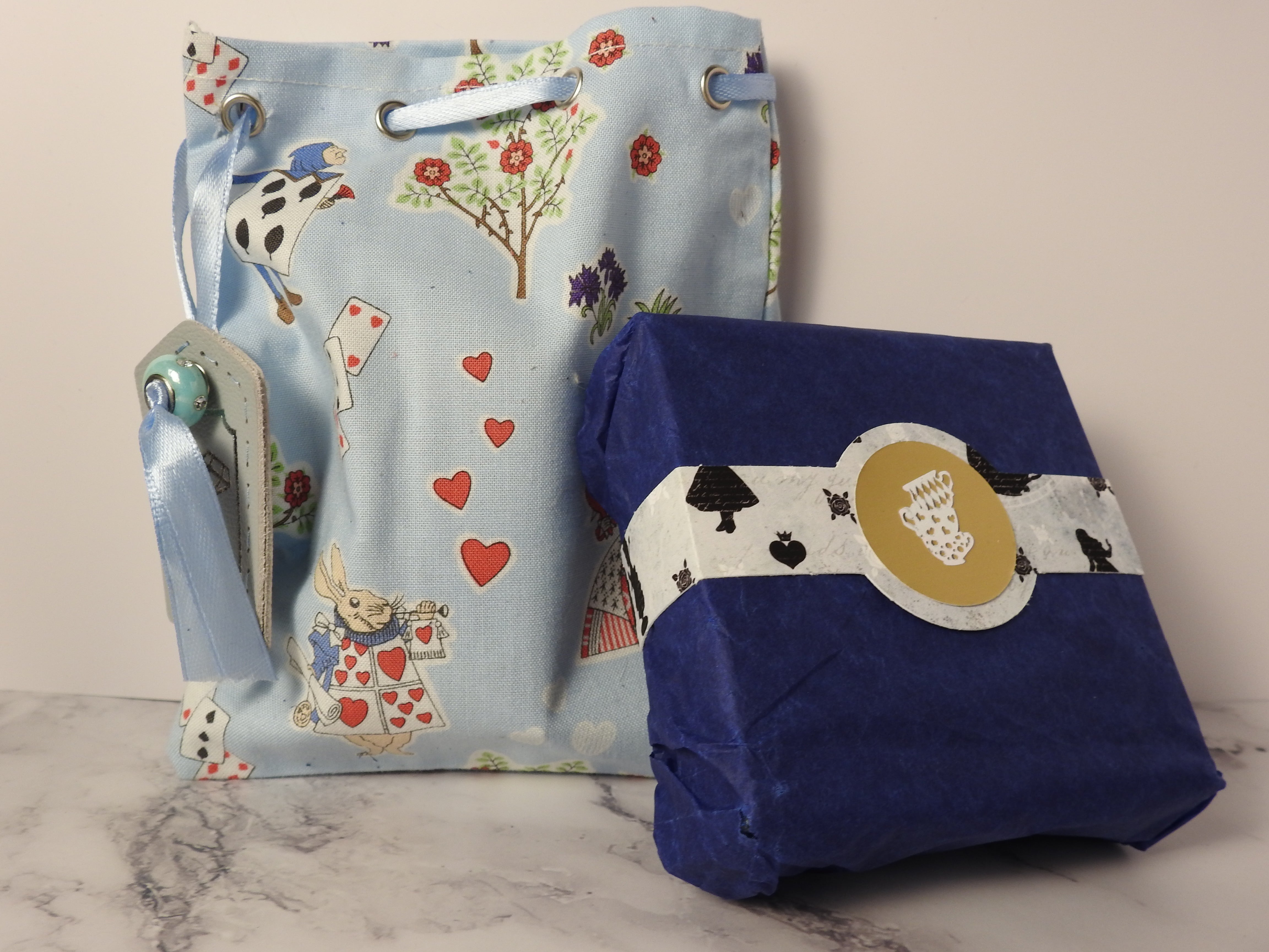 close up shot of a cotton bag and cardboard box with belly band. The cotton bag has an alice in wonderland printin blue and a leather tag, in the centre of the alice patterned belly band is a silver foil emblem of stacked teacups.