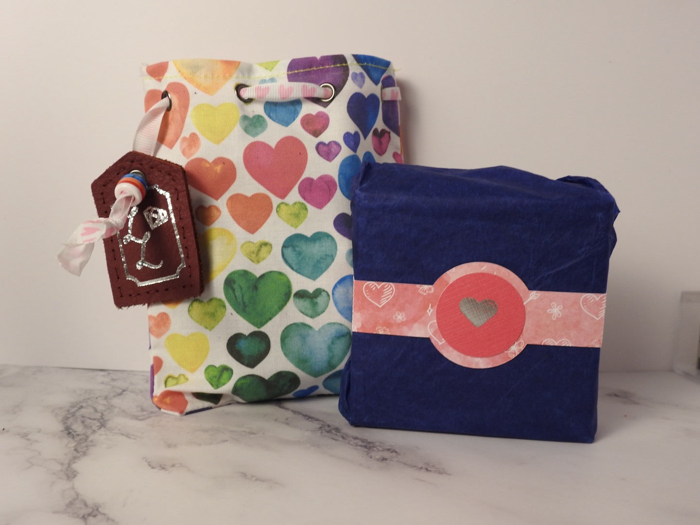 close up shot of a cotton bag and cardboard box with belly band. The cotton bag has valentines heart pattern in many colours and a leather tag, in the centre of the heart patterned belly band is a silver foil emblem of a valentine heart.