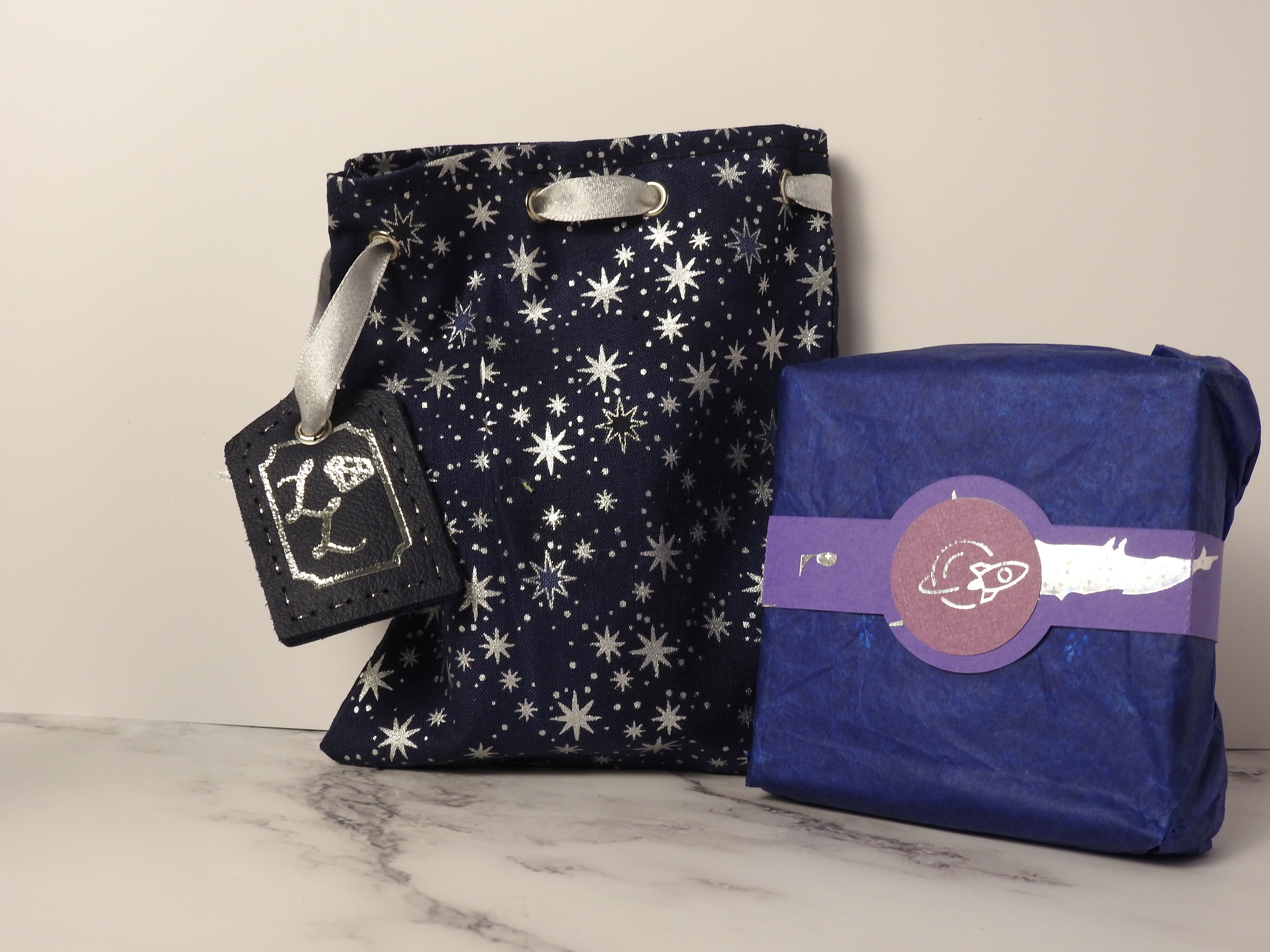 close up shot of a cotton bag and cardboard box with belly band. The cotton bag has starry pattern and a leather tag, in the centre of the purple belly band is a silver foil emblem of a rocket.