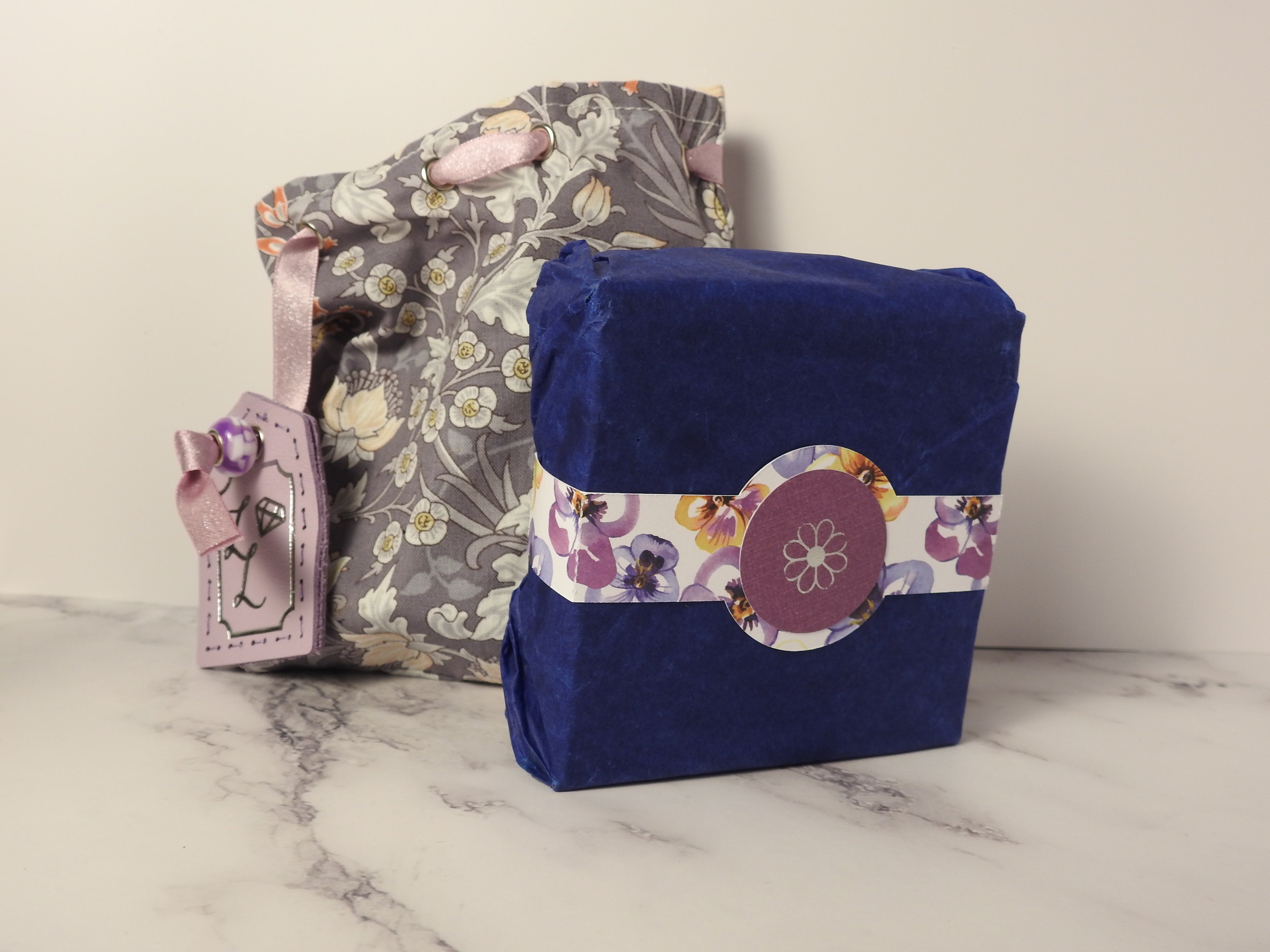 close up shot of a cotton bag and cardboard box with belly band. The cotton bag has floral patterns and a leather tag, in the centre of the floral patterned belly band is a silver foil emblem of a flower.