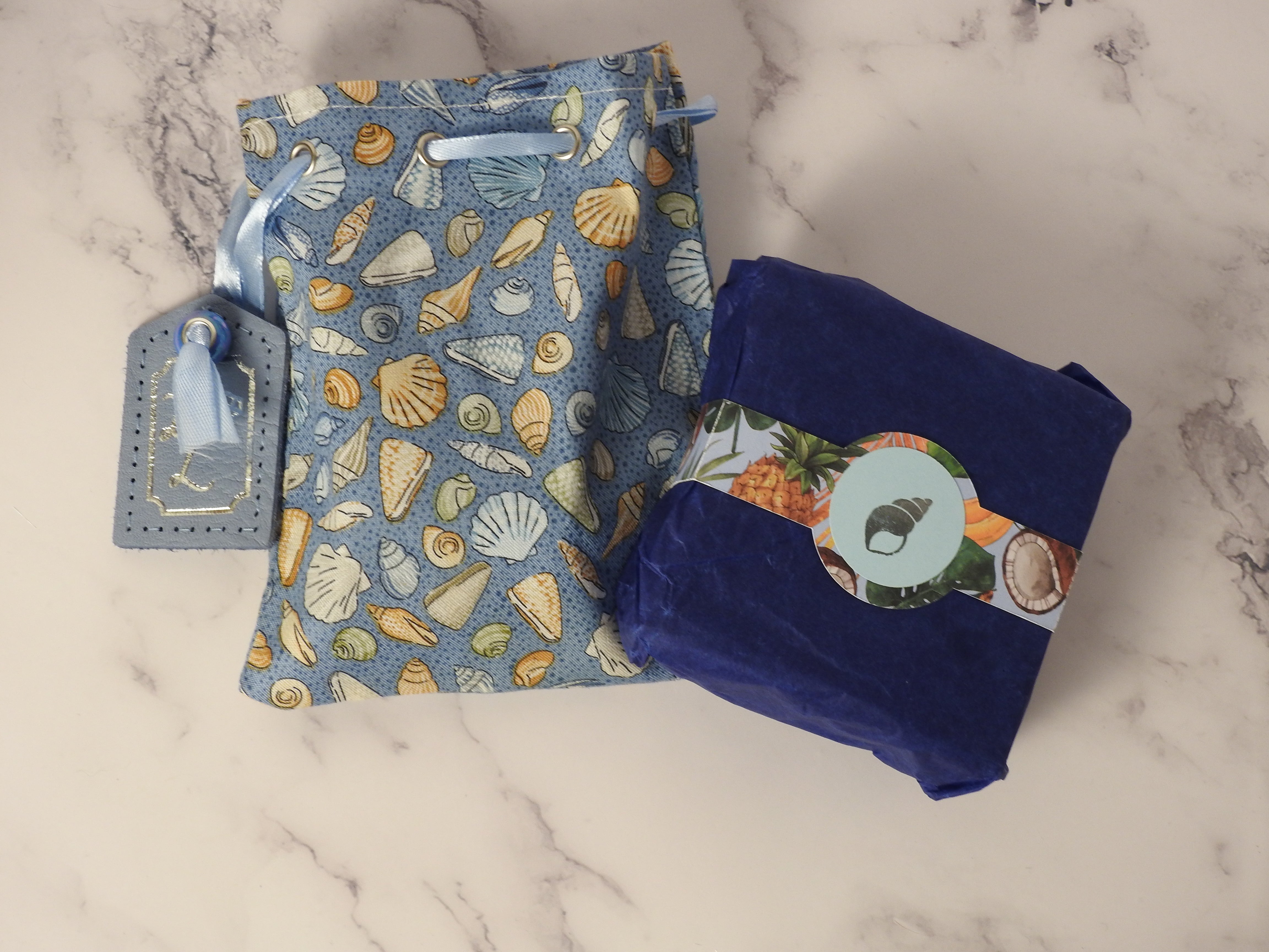 close up shot of a cotton bag and cardboard box with belly band. The cotton bag has seashell pattern in blue and a leather tag, in the centre of the summer patterned belly band is a silver foil emblem of a seashell.