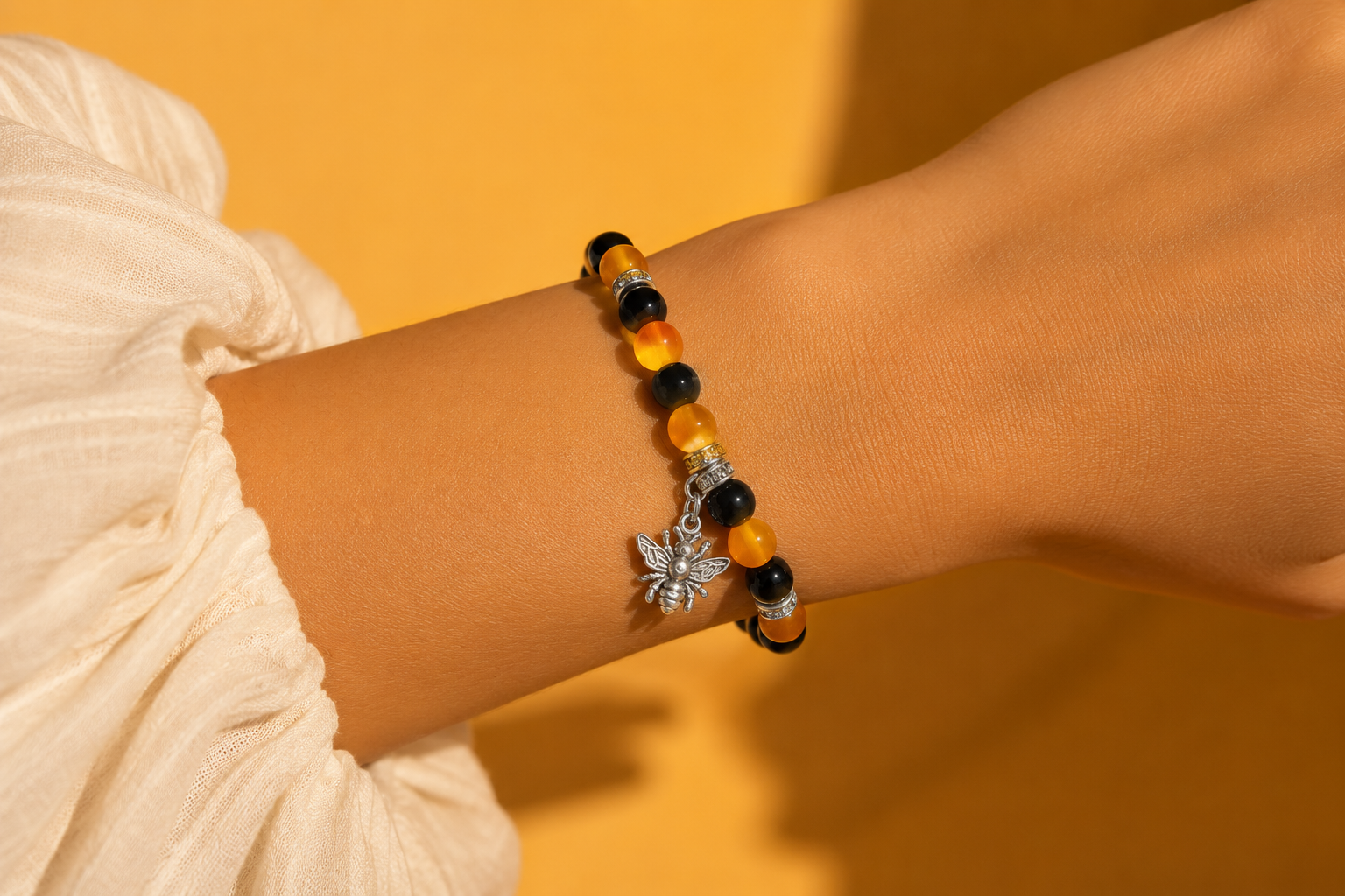 A close-up of a person's wrist wearing a beaded bracelet with black and orange beads, a sterling silver bee charm, and small silver spacer beads, set against a warm yellow background.