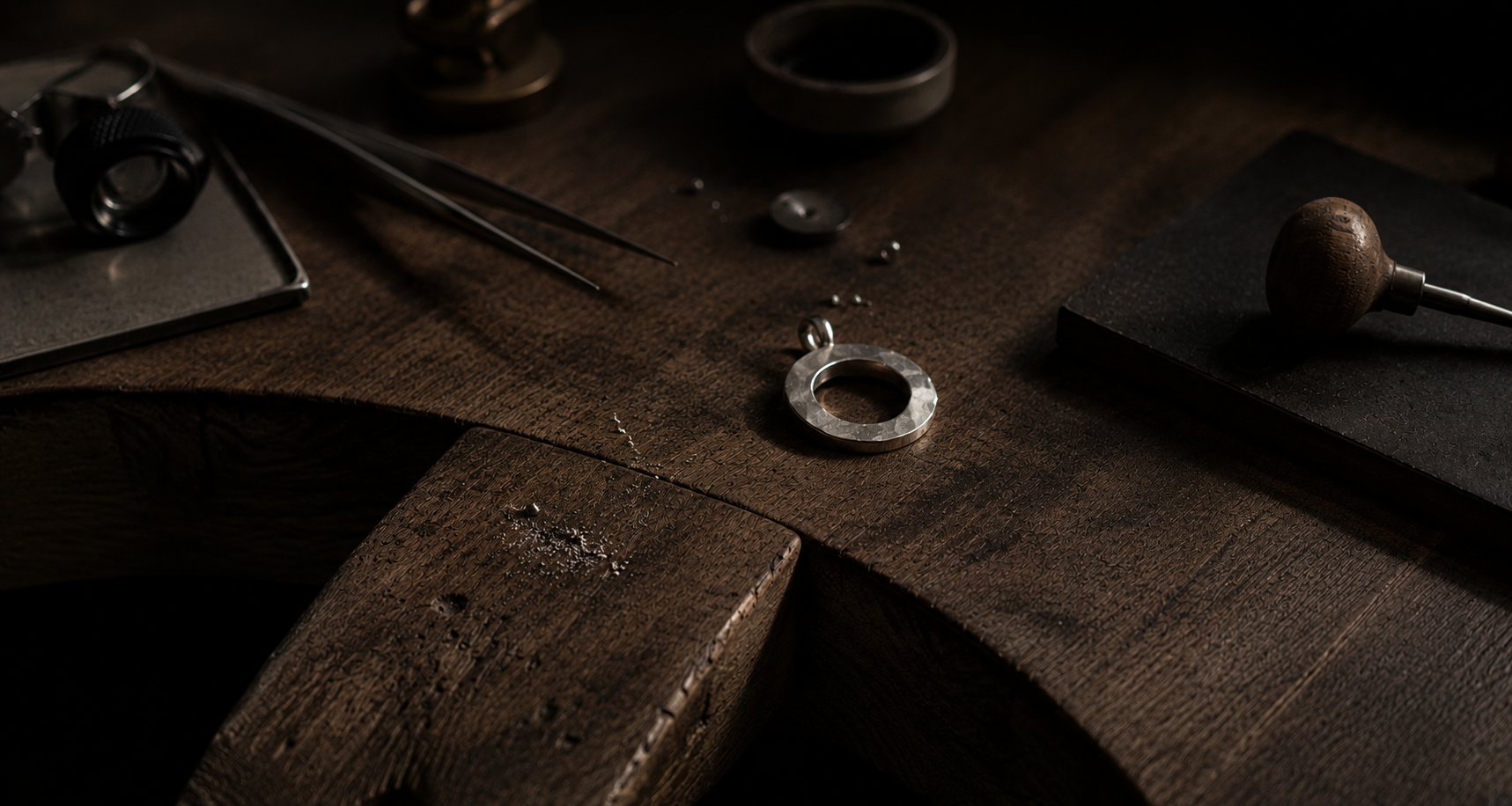 A silver ring blank with a small bail is displayed on a dark wooden workbench surrounded by jewelry-making tools and materials.