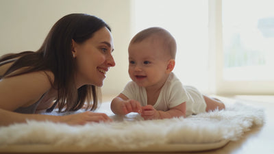 mother laid besides her baby on a wool rug, smiling at each other.