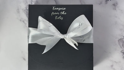 video of Lynne Le packing being opened. The outer package is a box tied with a white bow which is printed with "Lynne Lee" repeatedly in silver. At the top of the box is "treasure from the tides" debossed in silver foil. Inside the box is a cotton fabric bag in blue with seashell patterns, tied with a blue ribbon, a glass bead and leather tag. There is also a tiny bouquet of dried blue flowers. The cotton bag is then opened to reveal a box covered with blue tissue paper wrapped in a summer themed belly band