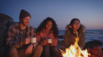 friends sat at night time in front of a campfire, the ocean in the background.